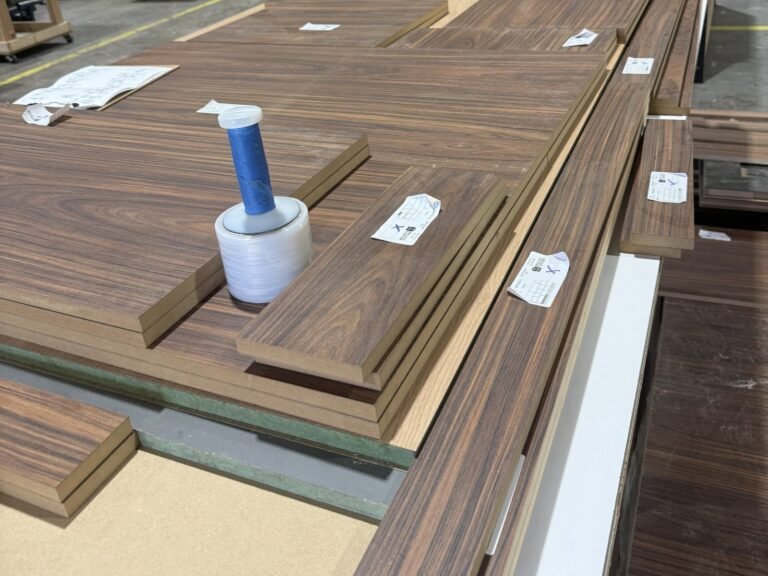 Edge-banded wood-grain cabinet parts with part labels stacked on a shop table, beside a printed parts sheet