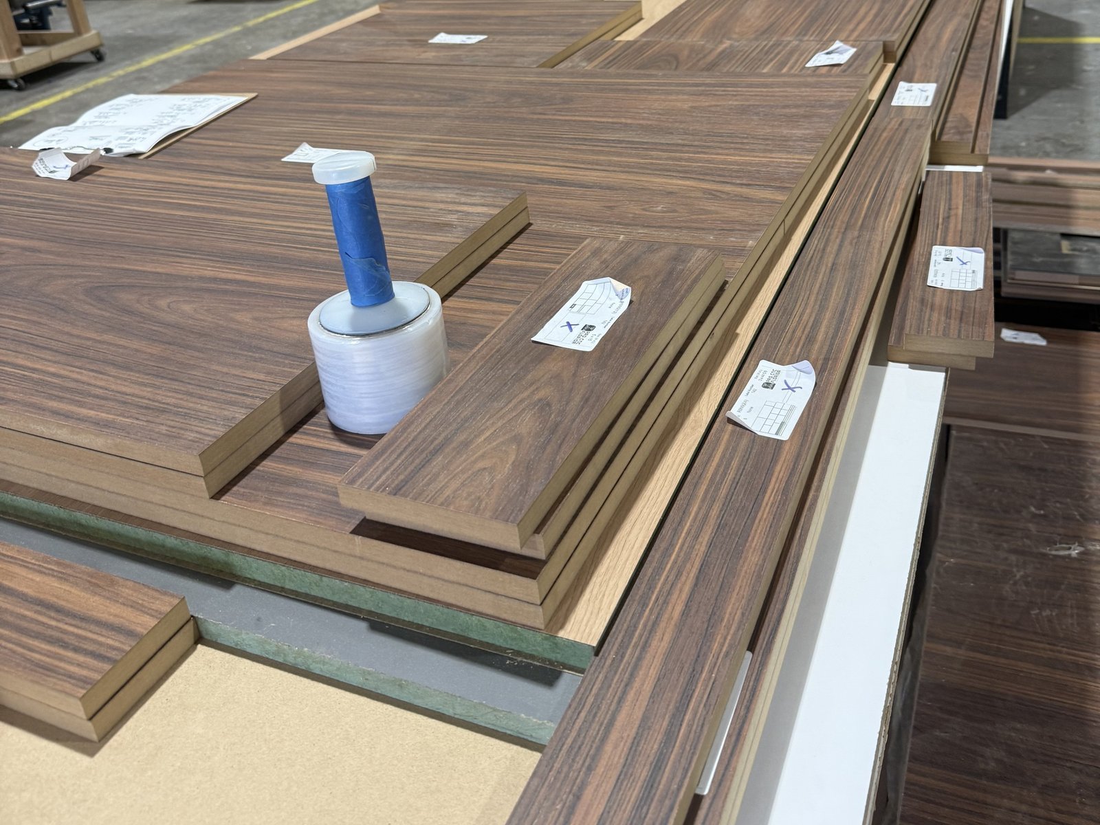 Edge-banded wood-grain cabinet parts with part labels stacked on a shop table, beside a printed parts sheet